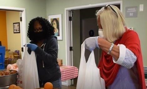 Two woman, one with a black sweater and another with a red and white sweater, are wearing blue gloves and sorting food items into shopping bags