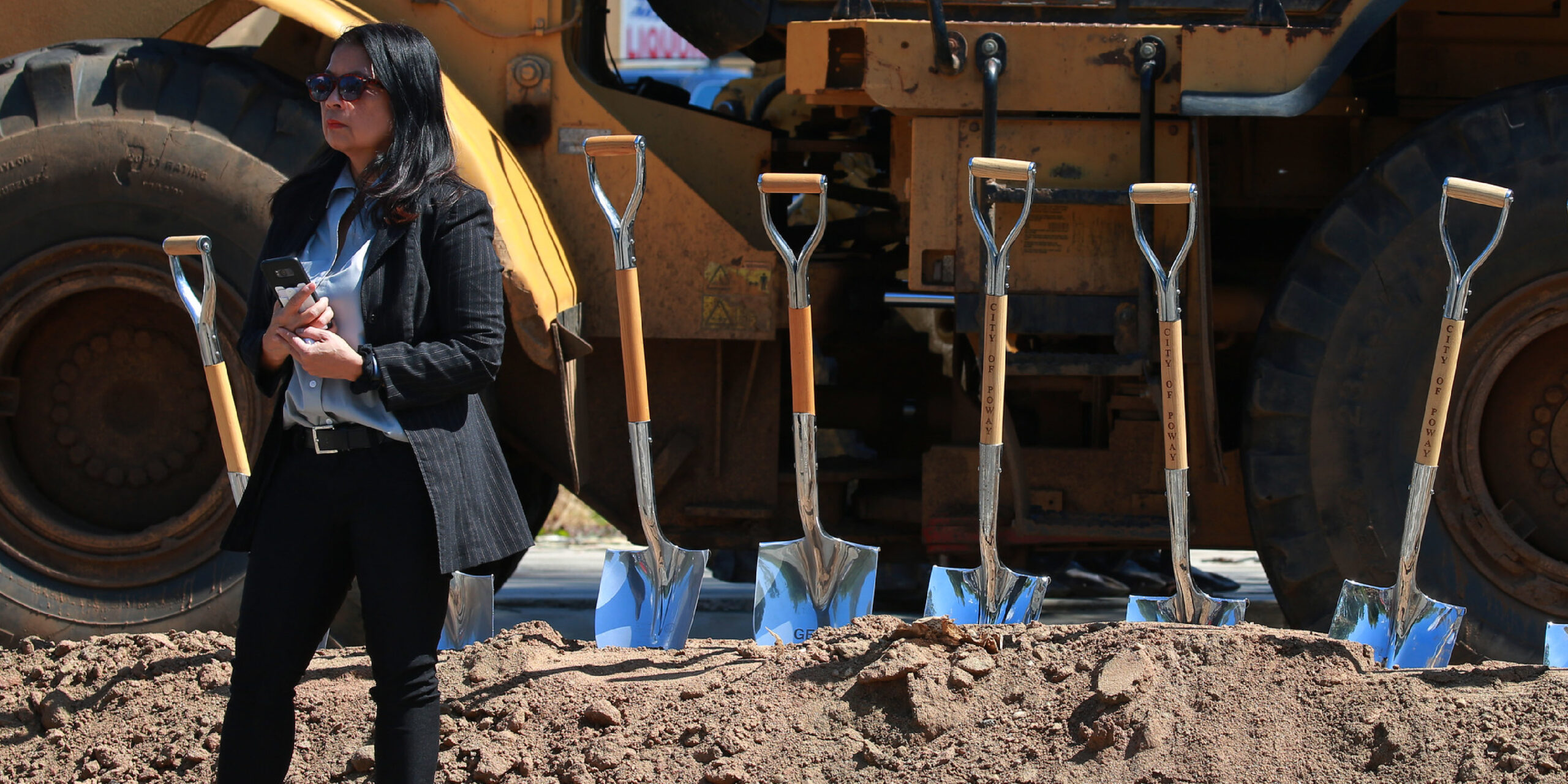 Amelita wearing a suit and sunglasses, standing outside in front of a small hill of dirt, with blue shovels lined up